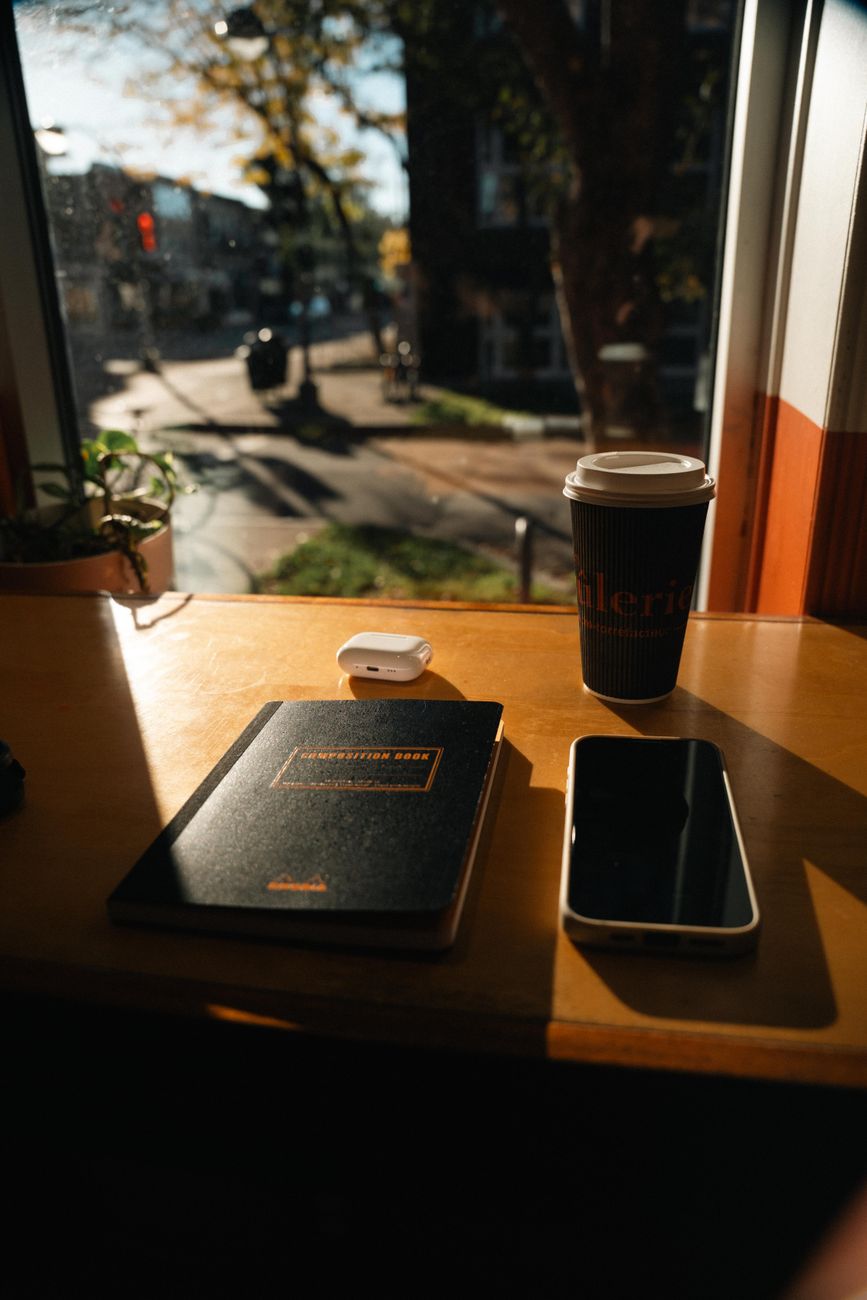 Notesbog, mobiltelefon og en kop kaffe på et træbord ved et vindue mod gaden